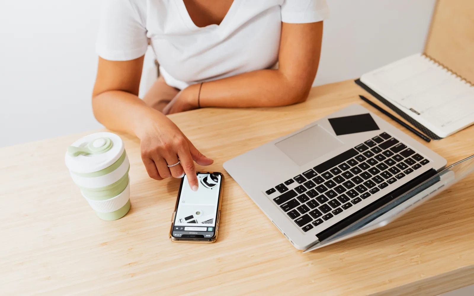 A person using a smartphone beside a laptop and notebook at a desk