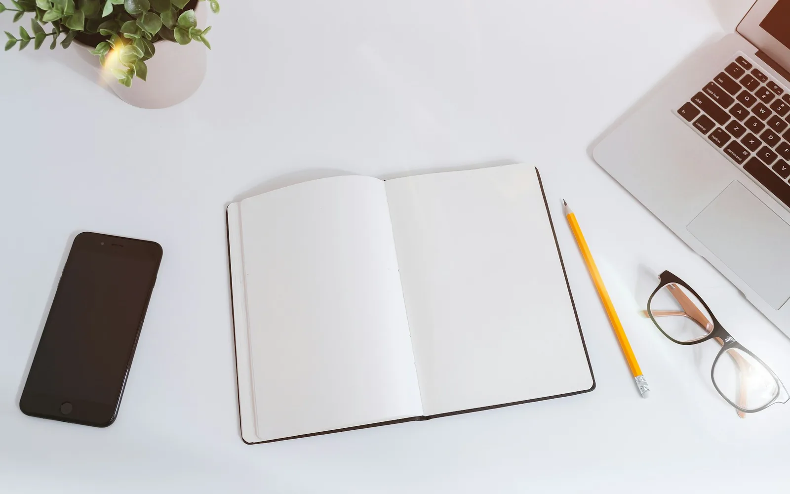An open notebook, pencil and glasses beside a laptop on a white desk